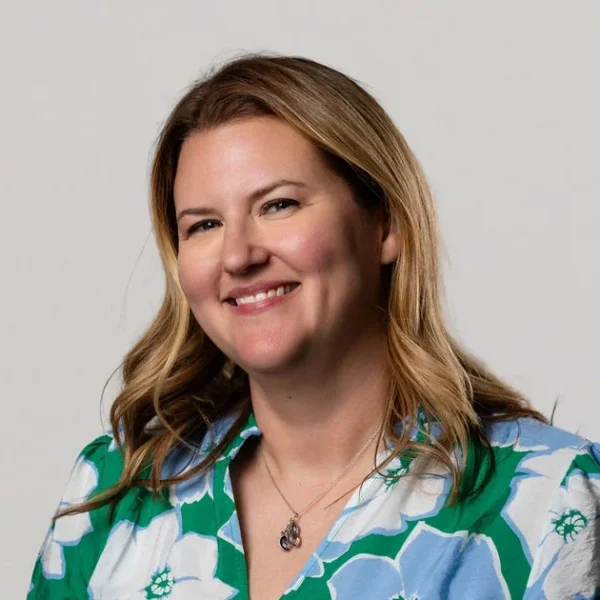 Woman with long blonde hair wearing a green blouse with large blue and white flowers smiles softly at the camera. Neutral background.